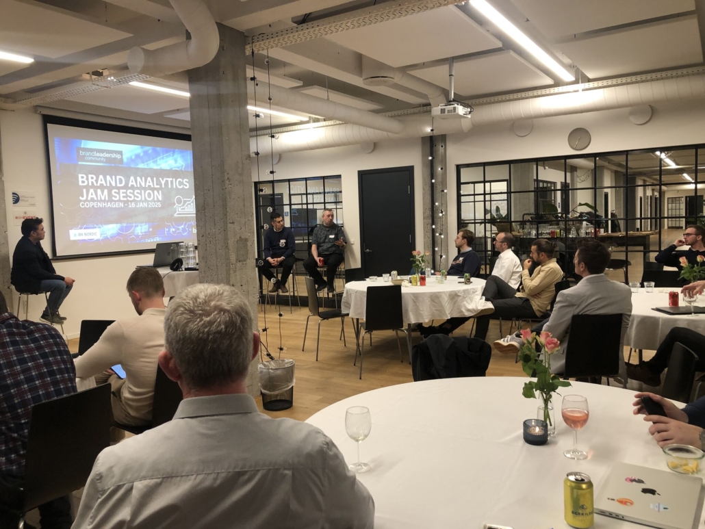 A group of professionals attending a Brand Analytics Jam Session in Copenhagen. The room features round tables with attendees, a projector screen displaying the event title, and speakers seated at the front.